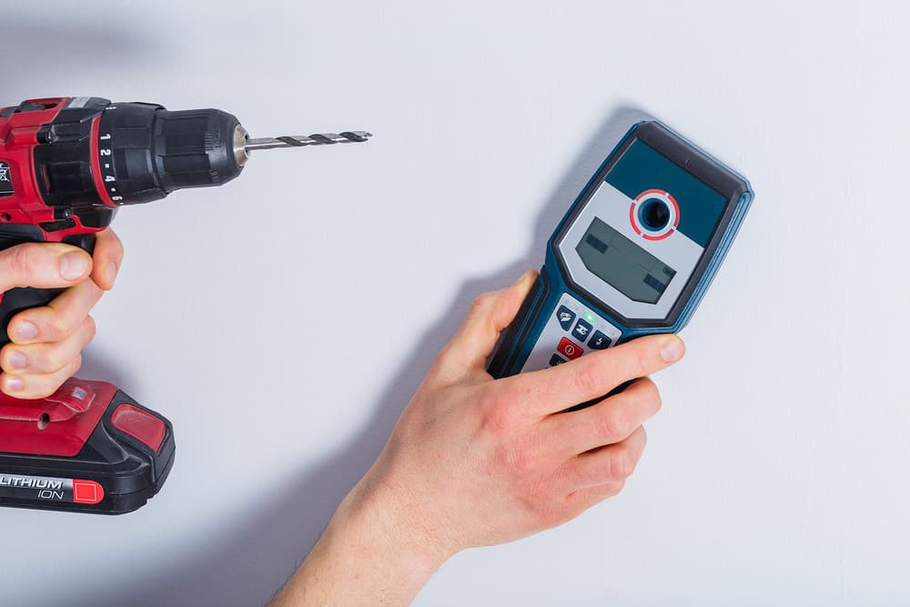Man scanning a wall with a stud finder while holding a drill in the other hand
