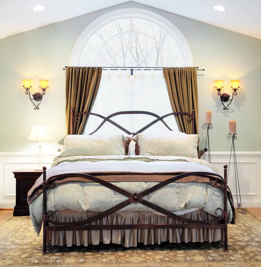 Interior of an upscale bedroom with arched window, vaulted ceiling, and ornate metal bed frame. Square format.