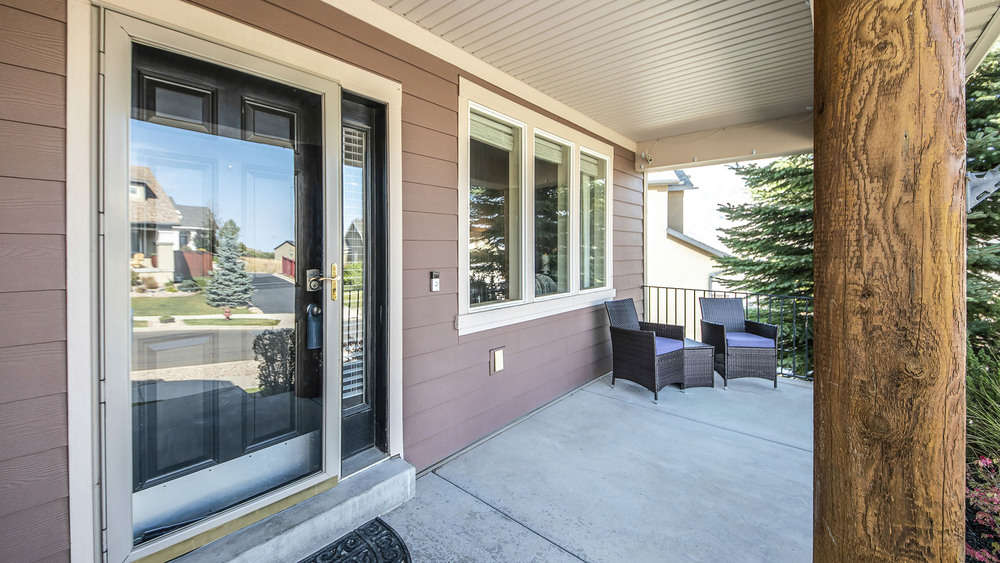 Pano Front porch of a house with engineered wood siding wall and log column post. Front exterior with storm door and side panel beside the window, and a set of chairs and table on the side.
