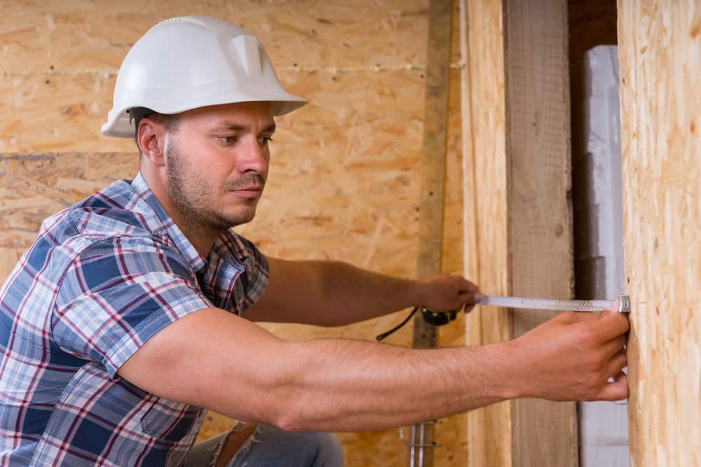 Construction Worker Builder Wearing White Hard Hat Measuring Width of Door Frame with Measuring Tape in Unfinished Home