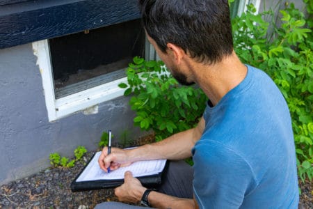 A close up and high angle view of a building inspector checking for defects near a basement window of a domestic home during an environmental quality exam.