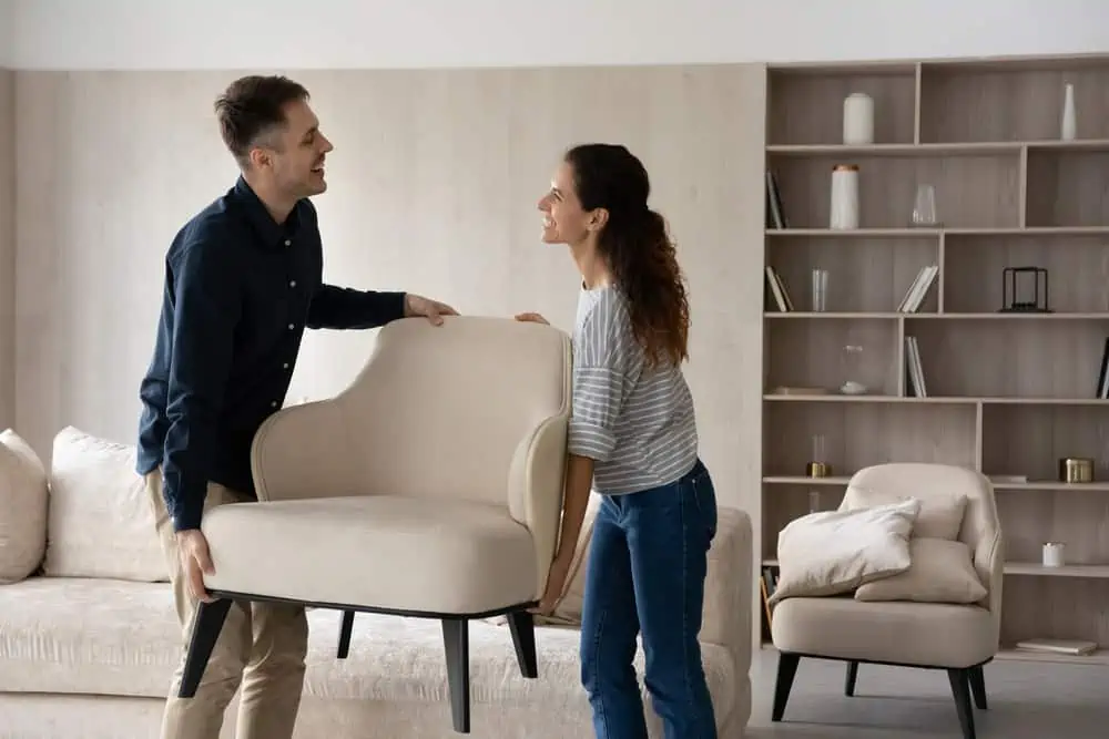 Happy hispanic young family couple homeowners holding armchair, enjoying renovating living room rearranging furniture together. Smiling spouses relocating on moving day into own dwelling.