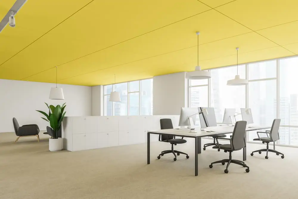 Corner of modern office with white walls, yellow ceiling, carpeted floor and two computer desks near white cabinets. Lounge area in background. 3d rendering