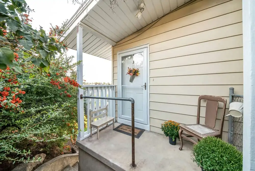 Front door exterior of a cottage style house with decorative flowers and plants. Small front porch of a house with a storm door and chairs at the front and a garden on the side.