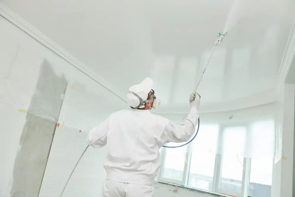 Painter worker with airless painting sprayer covering ceiling surface into white