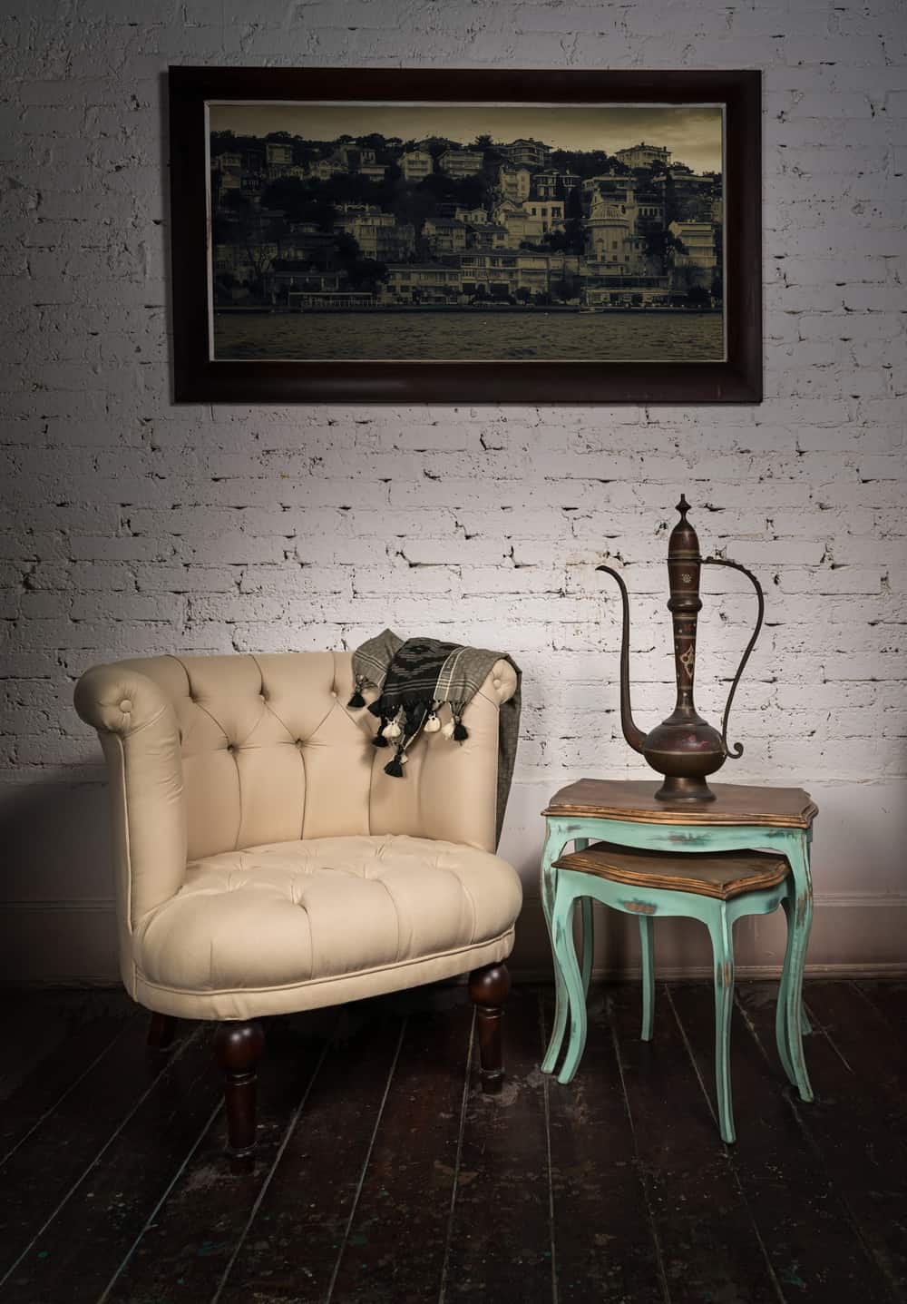 Vintage still life of old beige armchair, brass teapot, hanged framed painting and two small antique nested tables on dark brown wooden floor and white bricks wall in Studio
