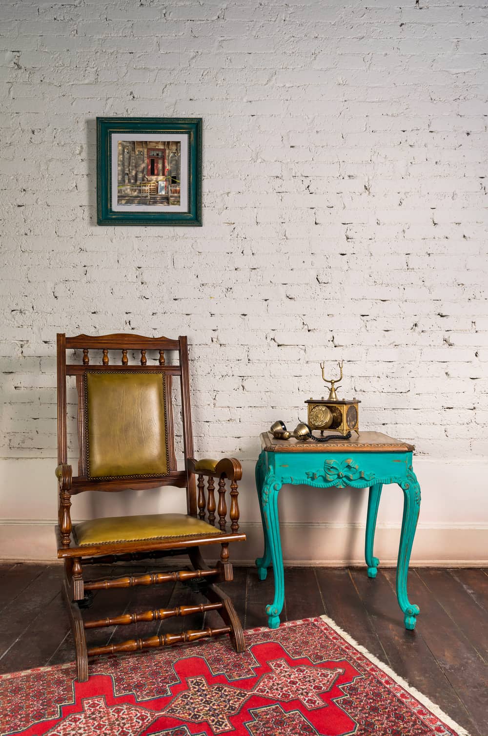 Classical wooden rocking chair, antique golden telephone set on top of green wooden vintage table and background of white bricks wall with hanged painting and parquet floor with red carpet