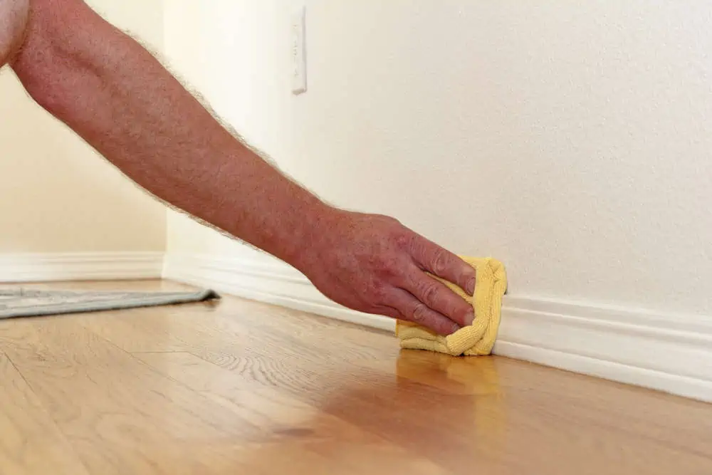 Male caucasian hand and arm seen wiping a folded yellow rag along the lower white wall trim near the wood floor to remove dust.