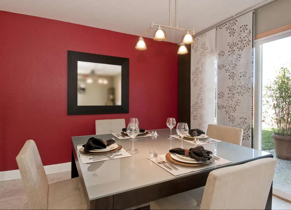modern dining room with glass top table, red wall with mirror, place settings, window shades, overhead lighting