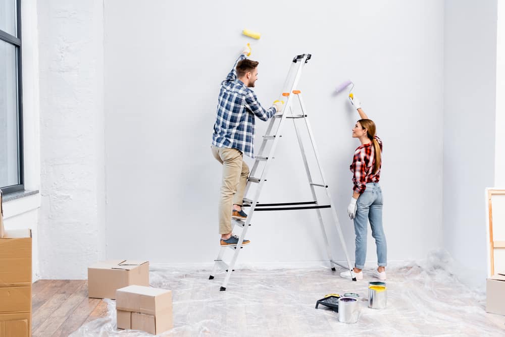 full length of smiling young couple with rollers painting wall and looking at each other near ladder at home