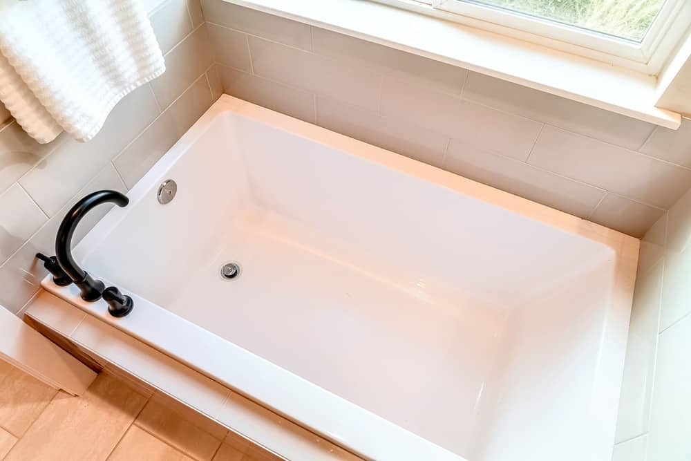 Built in rectangular bathtub with black gooseneck faucet inside home bathroom. Hanging towel and glass window can also be seen inside the room.