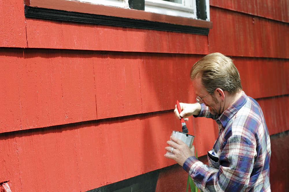 Mature male dipping a paintbrush in a can of black paint in order to touch up trim around the windows of his house on a sunny day.