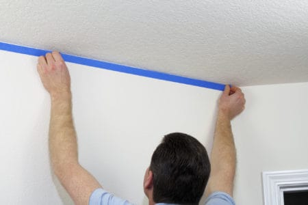 Man preparing to paint ceiling by masking off the wall beneath it with blue painter's tape.