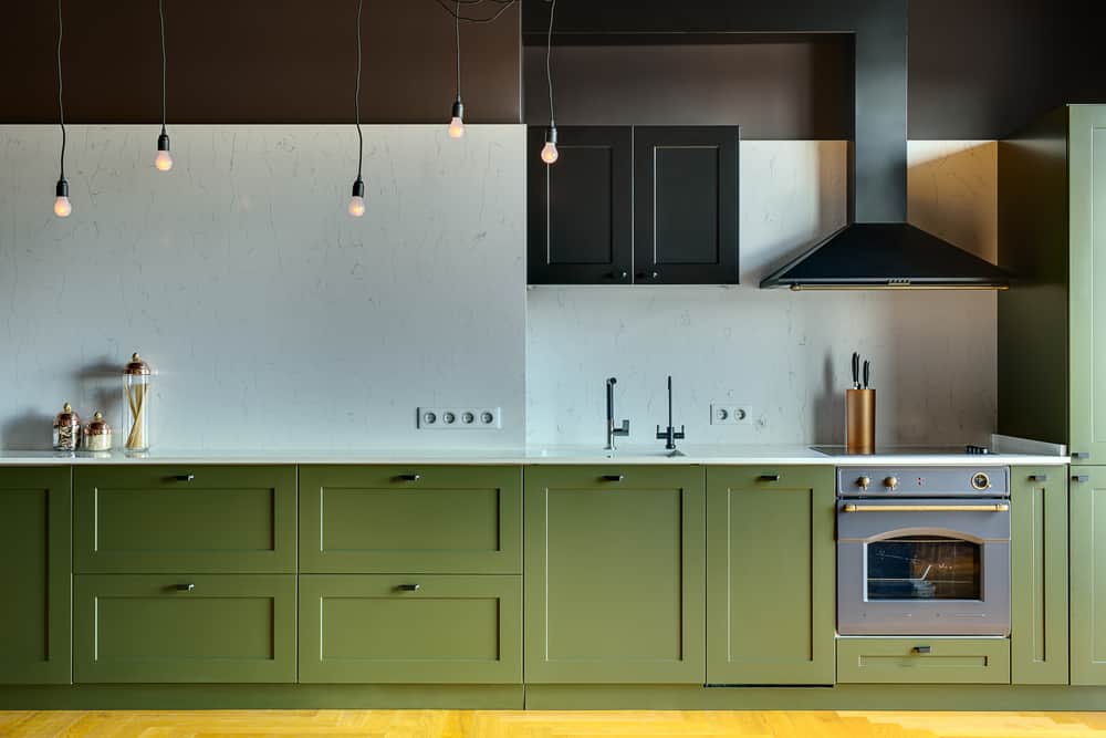 Kitchen in a modern style with a light tabletop with sink, cooker, oven, kitchen accessories. Under tabletop there are green drawers. Over tabletop there is kitchen hood, cupboard and glowing lamps.