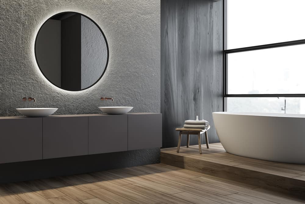 Corner of panoramic bathroom with concrete and gray walls, wooden floor, double sink standing on gray countertop with round mirror above it and comfortable bathtub. 3d rendering
