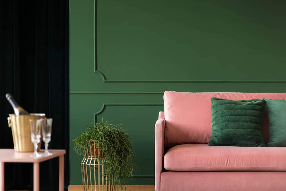 Flower next to pink couch with emerald pillows in dark green living room interior,copy space on empty wall