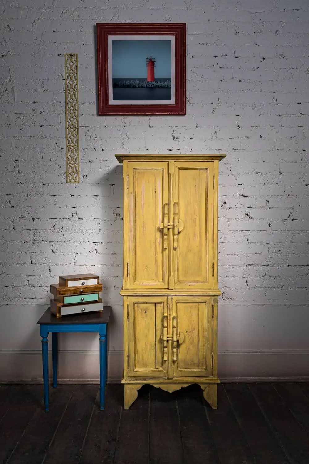 Still Life of vintage yellow cupboard, framed painting, small drawers unit and vintage small table on dark wooden floor and white bricks wall in Studio