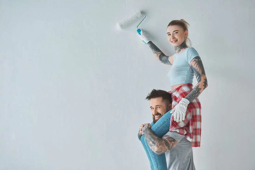 Smiling woman with roll paint brush sitting on boyfriends shoulders while painting wall at new home