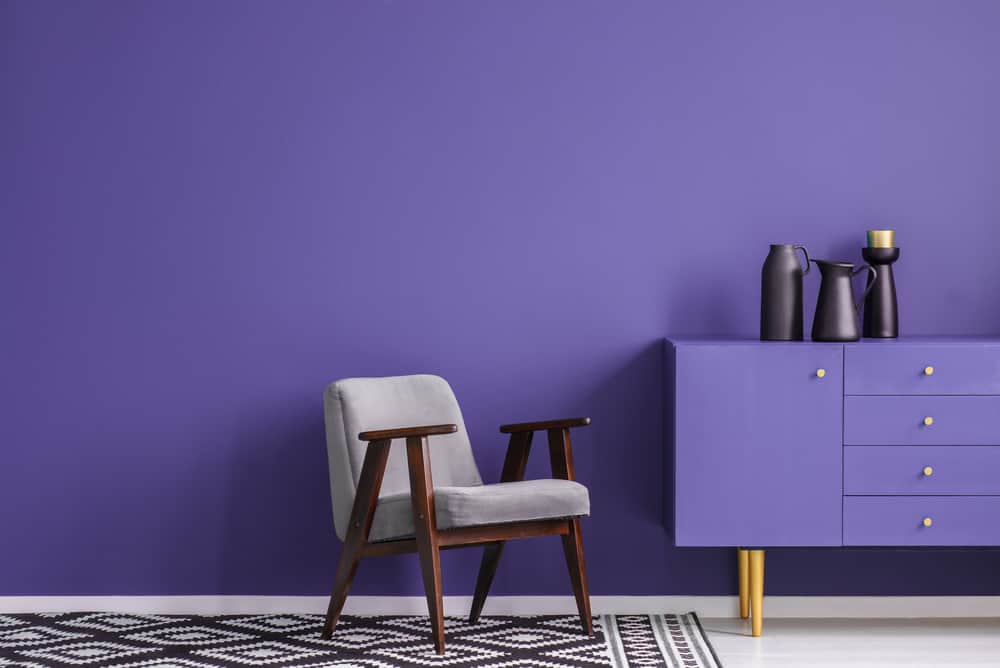 Grey armchair on patterned carpet next to violet cabinet with black vase in living room interior with copy space