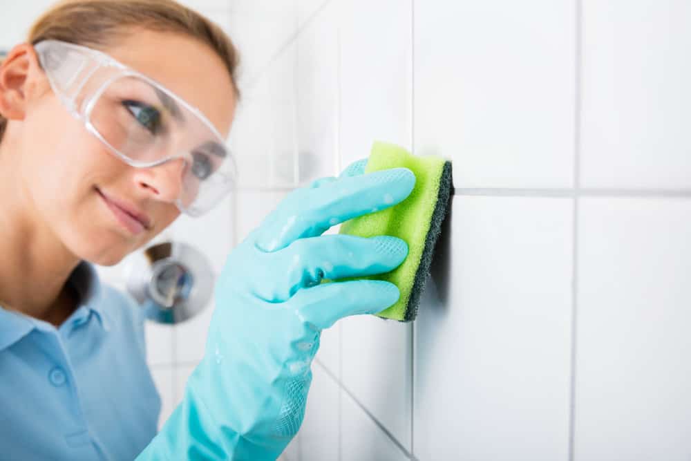 Young Woman With Protective Eyewear Cleaning The White Tile Of The Wall Using Sponge