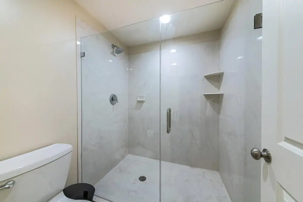 Interior of a bathroom with frameless shower stall. There is a white door on the right across the toilet beside the shower stall with glass enclosure and marble surround wall.