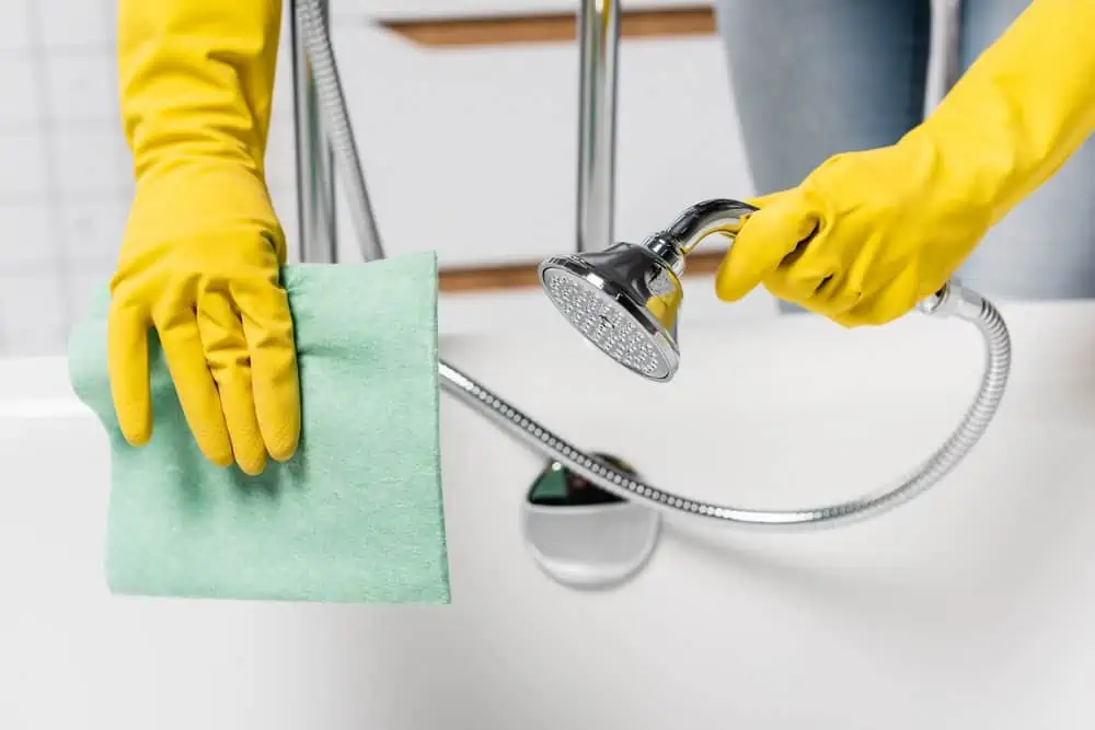 Female hands in yellow gloves holding shower head and cleaning the bathtub with green clotth
