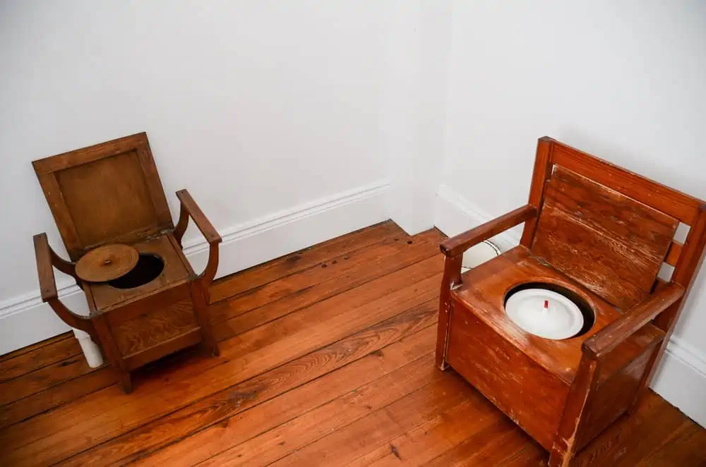 Old wooden dry composting toilets in room with white wall and wooden floor