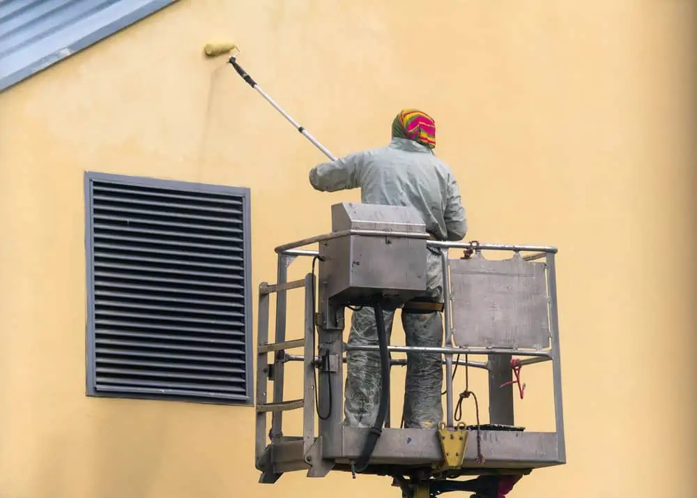 Man on lifting platform painting the house wall exterior yellow with roller