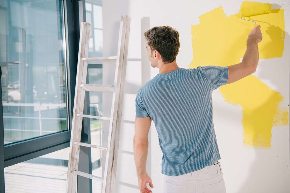 Man looking out the window while painting the house interior with yellow paint