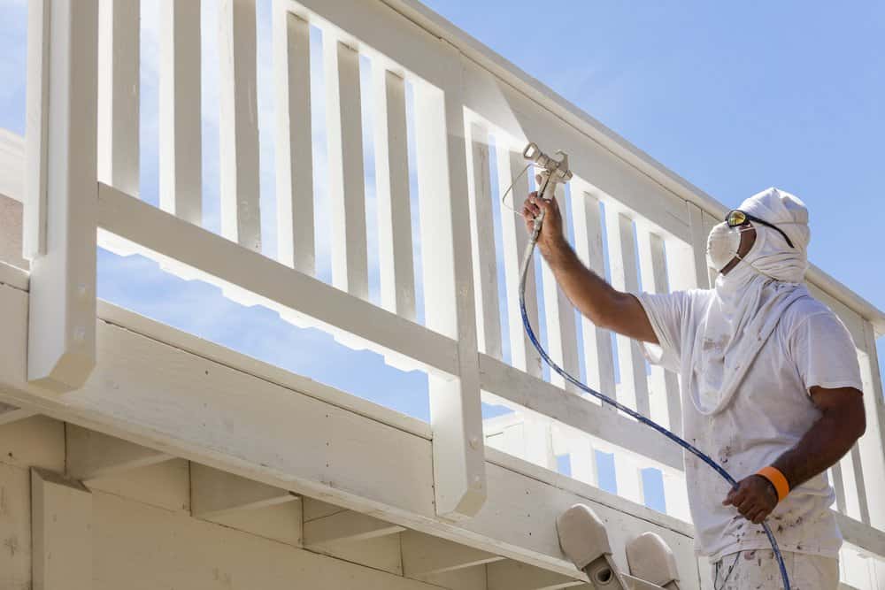 Painter wearing facial protection painting house deck white using spray gun