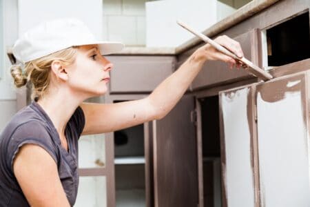 Woman wearing white cap holding paint brush and painting kitchen cabinets